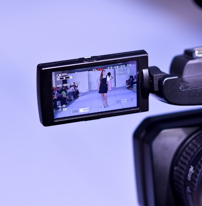 A video camera display screen shows a runway model walking on a catwalk, surrounded by an audience. The camera is capturing the scene, with the model wearing a dark blue dress.