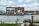 A large industrial-style building made of brick is situated beside a body of water. The building features numerous windows and a prominent mural of a cartoon figure riding a bicycle. The surroundings include some industrial equipment and a small boat docked nearby. The sky is partly cloudy, creating a calm atmosphere.