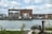 A large industrial-style building made of brick is situated beside a body of water. The building features numerous windows and a prominent mural of a cartoon figure riding a bicycle. The surroundings include some industrial equipment and a small boat docked nearby. The sky is partly cloudy, creating a calm atmosphere.