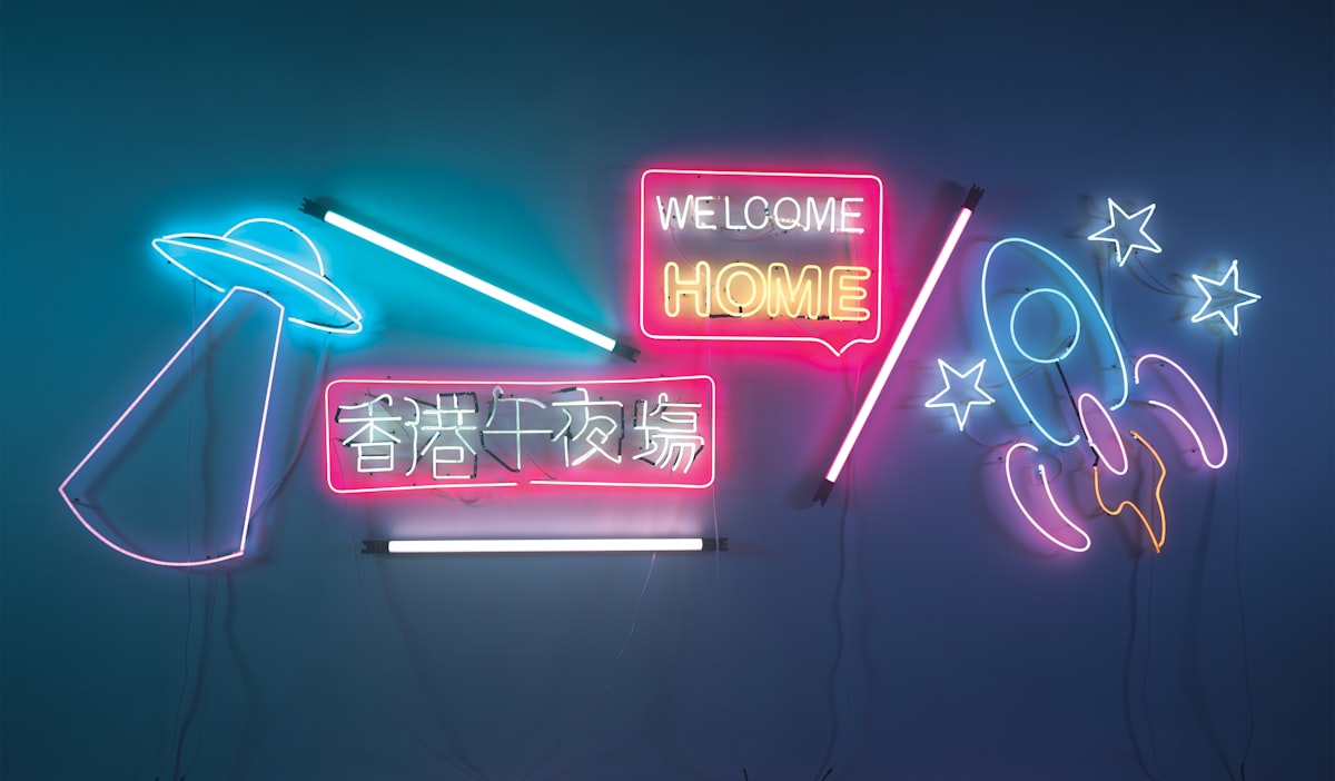 Neon signs on a wall: a flying saucer with a beam of light, a rocket, stars and a pink sign reading WELCOME HOME