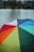 A vibrant, multicolored umbrella with sections in red, orange, yellow, green, teal, blue, and purple, covered in raindrops. It is set against a blurred background of a lake or river, with indistinct dark outlines of trees and cloudy skies.