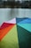 A vibrant, multicolored umbrella with sections in red, orange, yellow, green, teal, blue, and purple, covered in raindrops. It is set against a blurred background of a lake or river, with indistinct dark outlines of trees and cloudy skies.