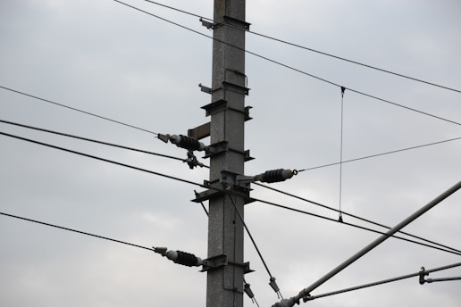 A utility pole is depicted with several electrical insulators and wires attached, set against a cloudy sky. The pole is composed of metal and features multiple cross-arms holding the insulators in place.