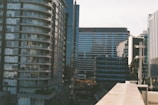 A cityscape featuring tall modern buildings with glass facades. The architecture includes a curved residential building on the left and commercial buildings with linear designs in the background. There is a blend of urban infrastructure and construction elements visible in the foreground.