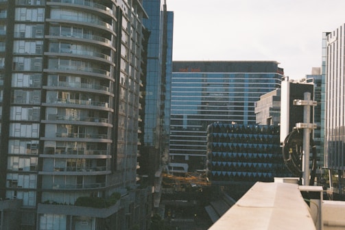A cityscape featuring tall modern buildings with glass facades. The architecture includes a curved residential building on the left and commercial buildings with linear designs in the background. There is a blend of urban infrastructure and construction elements visible in the foreground.