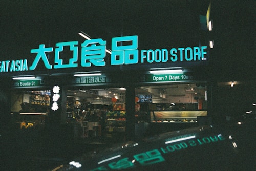 A storefront with bright neon signs displaying both English and Chinese text, indicating it's a food store open daily. The interior, partially visible, is stocked with various goods.