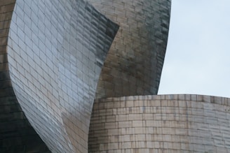 Curved metallic panels make up a complex architectural structure with overlapping surfaces. The smooth, reflective surface of the metal creates a dynamic visual pattern.