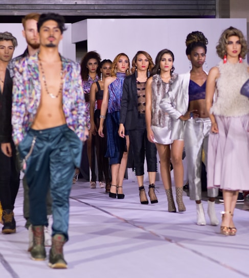 A diverse group of models walks confidently down a runway, showcasing a variety of fashion styles including metallic and patterned fabrics. The setting is a fashion show, and the models display an array of outfits ranging from casual to formal wear. Their expressions are serious and focused, reflecting the professional atmosphere of the event.