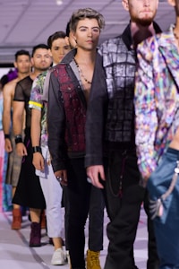 A group of male models walk down a runway during a fashion show. They are dressed in various colorful, trendy outfits with a focus on jackets and shirts. The lighting is bright, and the background is an indoor setting with ceiling panels and spotlights. The models appear focused and confident as they showcase the clothing items.