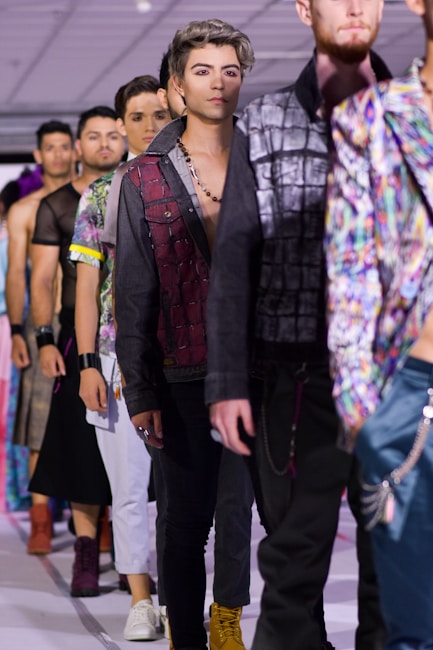 A group of male models walk down a runway during a fashion show. They are dressed in various colorful, trendy outfits with a focus on jackets and shirts. The lighting is bright, and the background is an indoor setting with ceiling panels and spotlights. The models appear focused and confident as they showcase the clothing items.