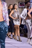 A group of models walking down a runway, with each wearing stylish outfits and high-heeled shoes. The models are arranged in a line, showcasing diverse fashion designs. The background includes a framed photo and spectators seated along the runway.
