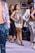 A group of models walking down a runway, with each wearing stylish outfits and high-heeled shoes. The models are arranged in a line, showcasing diverse fashion designs. The background includes a framed photo and spectators seated along the runway.