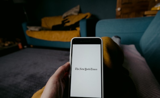 A person is holding a smartphone displaying The New York Times logo on its screen. The background features a cozy living room setting with a mustard yellow blanket draped over a couch and a teal sofa nearby.