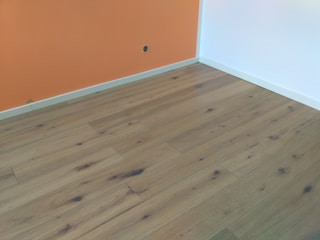 A room with wooden flooring, featuring natural grain patterns and knots, bordered by an orange and white wall with a small power outlet visible on the orange section.