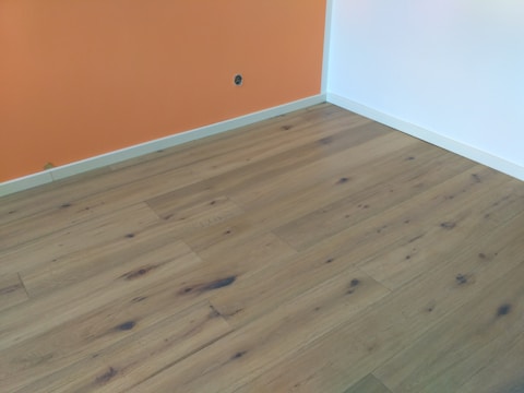 A room with wooden flooring, featuring natural grain patterns and knots, bordered by an orange and white wall with a small power outlet visible on the orange section.