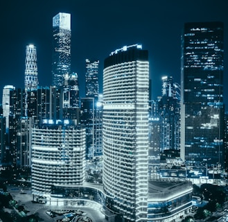 A cityscape at night featuring tall skyscrapers illuminated with bright lights. The skyline is dotted with various high-rise buildings, each showcasing modern architecture and design. The city appears vibrant and bustling, with a cool blue hue dominating the scene, accentuated by glowing windows and lights.