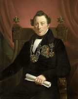 A portrait of a distinguished man seated on an ornate chair, wearing formal attire with various medals and decorations on his chest. He holds a rolled document in one hand and sits against a backdrop of a rich, red curtain. The chair features decorative elements, including two five-pointed stars at the top.