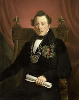 A portrait of a distinguished man seated on an ornate chair, wearing formal attire with various medals and decorations on his chest. He holds a rolled document in one hand and sits against a backdrop of a rich, red curtain. The chair features decorative elements, including two five-pointed stars at the top.