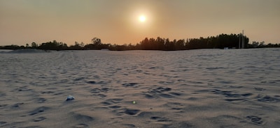 A vast sandy landscape stretches out with gentle undulations, capturing the texture of the sand beneath a setting sun. The horizon is lined with a silhouette of trees and a few sparse structures. The sky is a soft gradient of orange and yellow, enhancing the serene atmosphere.