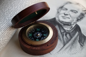 A wooden compass with a brass rim lies open next to a page of text and a black-and-white sketch of a bearded man. The compass needle points north, and the intricate design on its face is clearly visible.