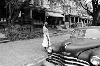 grayscale photo of woman in long sleeve dress standing beside car