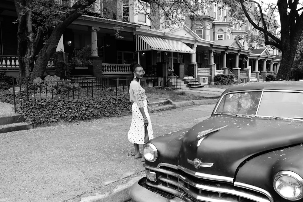 grayscale photo of woman in long sleeve dress standing beside car