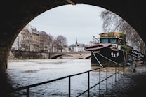 A vintage boat is moored along a flooded riverside in a European city. The scene is framed by the arch of a bridge, with buildings and leafless trees lining the street above the river. The architecture is classic with apartments featuring wrought iron balconies. The overcast sky and reflections in the water add a serene yet somber atmosphere.