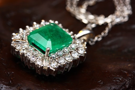 An emerald gemstone is set at the center of an intricate silver pendant surrounded by sparkling diamonds. The pendant's chain, also silver, is delicately intertwined. The background features a dark, textured surface that contrasts with the shimmering jewelry.