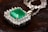 An emerald gemstone is set at the center of an intricate silver pendant surrounded by sparkling diamonds. The pendant's chain, also silver, is delicately intertwined. The background features a dark, textured surface that contrasts with the shimmering jewelry.