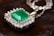 An emerald gemstone is set at the center of an intricate silver pendant surrounded by sparkling diamonds. The pendant's chain, also silver, is delicately intertwined. The background features a dark, textured surface that contrasts with the shimmering jewelry.