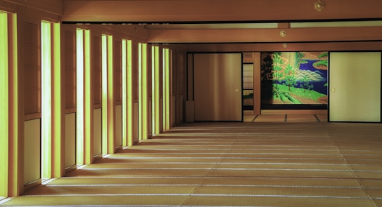 A traditional Japanese room with sliding doors, wooden beams, and tatami mat flooring. The walls have vertical paper panels that allow natural light to stream in, creating a serene and well-lit atmosphere. There is a vibrant mural on one of the sliding doors depicting a natural scene with trees and a body of water.
