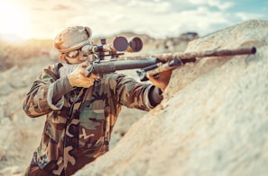 A person in camouflage clothing aims a rifle with a scope, positioned against a rocky surface. The individual wears a cap and protective eyewear, suggesting a tactical or hunting scenario. The environment appears to be outdoors, likely in a rugged or mountainous area with overcast or diffused lighting.