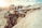 A person in camouflage clothing aims a rifle with a scope, positioned against a rocky surface. The individual wears a cap and protective eyewear, suggesting a tactical or hunting scenario. The environment appears to be outdoors, likely in a rugged or mountainous area with overcast or diffused lighting.