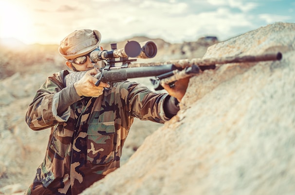 A person in camouflage clothing aims a rifle with a scope, positioned against a rocky surface. The individual wears a cap and protective eyewear, suggesting a tactical or hunting scenario. The environment appears to be outdoors, likely in a rugged or mountainous area with overcast or diffused lighting.