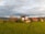 A picturesque countryside scene features a large, white building with a red roof situated on a lush green hill. The landscape includes several other rustic buildings, a few tall trees, and a winding dirt path leading towards the structures. The sky is overcast, casting a soft, diffused light over the scene.