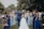 A wedding celebration with a bride and groom walking down an aisle outdoors, surrounded by a cheering crowd. The bridesmaids are wearing varying shades of blue dresses, and the atmosphere is festive. People are holding bouquets and capturing moments on their phones, while a child sits on a chair observing the scene.