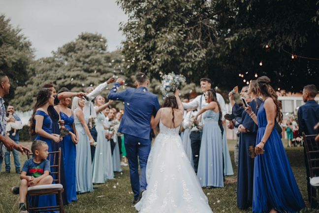 A wedding celebration with a bride and groom walking down an aisle outdoors, surrounded by a cheering crowd. The bridesmaids are wearing varying shades of blue dresses, and the atmosphere is festive. People are holding bouquets and capturing moments on their phones, while a child sits on a chair observing the scene.
