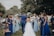 A wedding celebration with a bride and groom walking down an aisle outdoors, surrounded by a cheering crowd. The bridesmaids are wearing varying shades of blue dresses, and the atmosphere is festive. People are holding bouquets and capturing moments on their phones, while a child sits on a chair observing the scene.