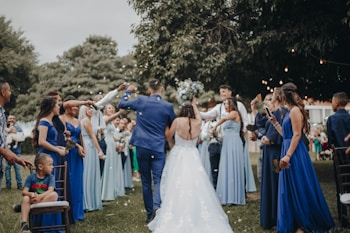 A wedding celebration with a bride and groom walking down an aisle outdoors, surrounded by a cheering crowd. The bridesmaids are wearing varying shades of blue dresses, and the atmosphere is festive. People are holding bouquets and capturing moments on their phones, while a child sits on a chair observing the scene.