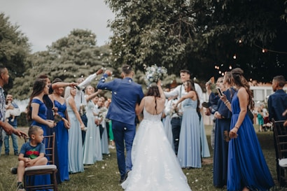 A wedding celebration with a bride and groom walking down an aisle outdoors, surrounded by a cheering crowd. The bridesmaids are wearing varying shades of blue dresses, and the atmosphere is festive. People are holding bouquets and capturing moments on their phones, while a child sits on a chair observing the scene.