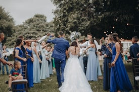 A wedding celebration with a bride and groom walking down an aisle outdoors, surrounded by a cheering crowd. The bridesmaids are wearing varying shades of blue dresses, and the atmosphere is festive. People are holding bouquets and capturing moments on their phones, while a child sits on a chair observing the scene.