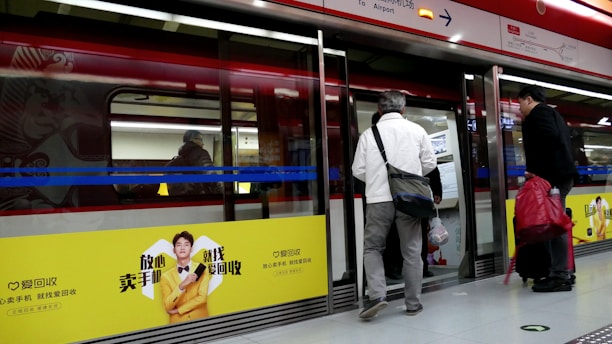 Passengers are waiting to board a metro train at a station. The platform doors are open, and several people with luggage are preparing to enter the train. A large advertisement with a bright yellow background is displayed on the train, featuring a person and text in Chinese.