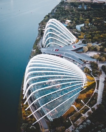 A large, modern greenhouse with glass panels and white structural beams arches over a lush garden. The structure is situated near a body of water, surrounded by vegetation and pathways. Its sleek, curved design integrates with the natural setting, reflecting the sky and providing a futuristic aesthetic.