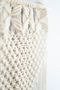 Intricate macramé pattern featuring woven and knotted cord designs, creating a textured and airy appearance. The focus is on the detailed craftsmanship of the knots against a soft, neutral background.