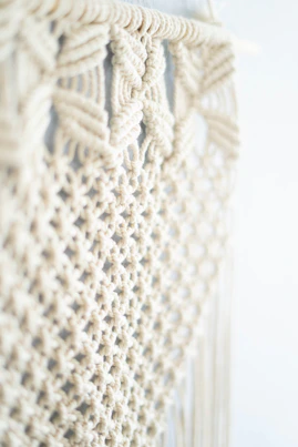 Close-up of hands weaving natural cotton macrame cords into an intricate wall hanging, surrounded by soft earthy tones.