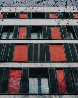 A modern building fa&ccedil;ade featuring a symmetrical arrangement of windows with dark shutters against a backdrop of red panels. The structure is complemented by shadows of tree branches cast on the walls, adding depth and texture to the scene.