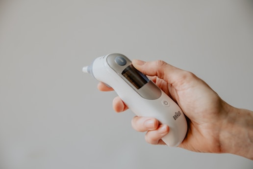 A hand is holding a digital infrared thermometer, which is primarily white with a gray display screen and a circular button. The background is a soft, neutral color that enhances the focus on the thermometer.