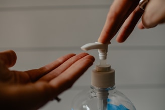 A close-up of a person's hands using a pump bottle to dispense a liquid, with one hand pressing the pump and the other hand open to receive the liquid. The background is softly blurred, with horizontal lines visible.