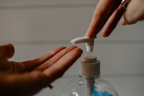 A close-up of a person's hands using a pump bottle to dispense a liquid, with one hand pressing the pump and the other hand open to receive the liquid. The background is softly blurred, with horizontal lines visible.