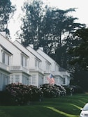 A row of white, two-story houses with gabled roofs is adorned by well-maintained gardens filled with hydrangea bushes. An American flag is prominently displayed in front of one house. Mature trees with dense foliage are visible in the background, creating a serene suburban setting. The lawn is neatly cut, and a car is partially visible on the right side of the image.